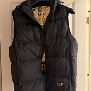 Svart dunväst från Fashion Black Gold - Säljer en svart dunväst från Black Gold med huva och dragkedja framtill. Insidan har ett coolt guldigt hexagonmönster som ger extra edge. Västen är quiltad och har en loose passform, perfekt att slänga över en hoodie när det är kallt ute.