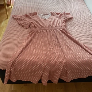 Rosa prickig kortklänning med volang - Supersöt rosa kortklänning med vita prickar och volangkant. Klänningen har korta ärmar, v-ringning och ett smockat midjeband som ger en snygg siluett. Perfekt för dig som gillar lekfulla och trendiga detaljer.