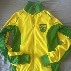 Gul och grön Brasil track jacket S - Gul och grön track jacket med Brasil-tryck fram och bak. Jackan har hög krage, ribbade muddar och dragkedja framtill. Snygga gröna paneler längs ärmar och sidor, samt Brasil-märke på bröstet. Perfekt för dig som älskar fotboll och vill sticka ut.
