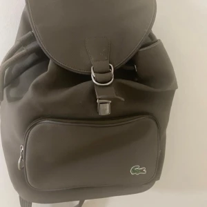 Brun ryggsäck från Lacoste - Snygg brun ryggsäck från Lacoste i slitstark polyester med klassisk krokodillogga framtill. Väskan har ett stort huvudfack med dragsko och lock, samt en praktisk ytterficka med dragkedja. Insidan är fodrad med grönt och beige randigt tyg. 