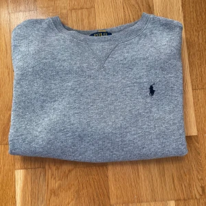 Grå sweatshirt från Polo Ralph Lauren - Klassisk grå sweatshirt från Polo Ralph Lauren med rund halsringning och broderad marinblå logga på bröstet. Tröjan är i mjuk bomullsmix och har ribbade muddar vid ärmslut och nederkant. Perfekt för en chill och stilren look. Tröjan är i bra skick och strlk 10-12 i junior vilket är typ 146-152, skriv vid minsta fråga!