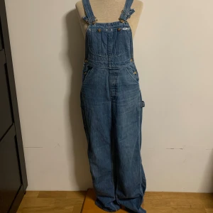 Blå denim hängselbyxor från Lee - Klassiska blå denim hängselbyxor från Lee med justerbara axelband. Snygga detaljer med Lee-logga och metallknappar.