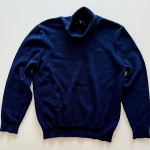 Mörkblå polotröja från Gant - Mysig mörkblå polotröja från Gant i mjuk ull. Tröjan har lång ärm, ribbade muddar och klassisk polokrage. Perfekt för kyliga dagar och enkel att matcha med jeans eller kostymbyxor.