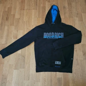 Svart hoodie från Hoodrich - Svart hoodie från Hoodrich med blått foder i huvan och blått tryck på bröstet och ryggen. Tröjan har snörning i huvan, stor magficka och coola broderade detaljer. Perfekt för dig som gillar streetwear och vill sticka ut lite extra. Pris kan diskuteras.