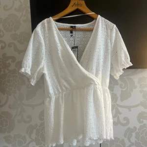 Vit omlottblus från Vero Moda - Superfin vit blus från Vero Moda med omlott och v-ringning. Blusen har korta puffärmar, broderade hålmönster och resår i midjan för en snygg passform. Perfekt till jeans eller kjol för en fräsch look.