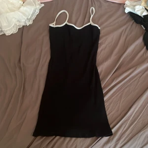 Svart kort klänning  - En svart, tight kortklänning med smala axelband och vita kontrastkanter längs urringningen. Klänningen har en enkel och stilren design som känns både trendig och klassisk. Den har u-ring vid ryggen