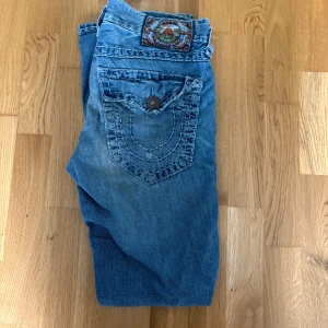 True Religion blå jeans, bootcut - Säljer ett par klassiska blå jeans från True Religion med tydliga kontrastsömmar och ikonisk bakficka. Jeansen har bootcut-modell och är tillverkade i bomull. Snygg tvättad look och detaljerad patch bak i midjan. measurements: 40cm-midja 21cm-benöppning 102cm-längd