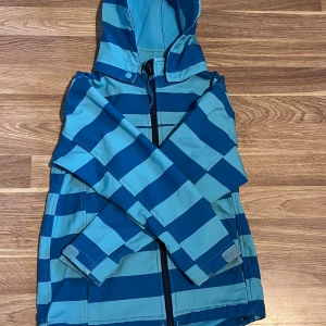 Blå randig vindjacka från Nico - Snygg vindjacka från Nico i blåa och turkosa ränder. Jackan har huva, dragkedja framtill och praktisk bröstficka med dragkedja. Materialet är vindtätt, vattentätt och andas, perfekt för blåsiga dagar. Justerbara ärmslut med kardborre. Perfekt för dig som gillar färg och funktion.