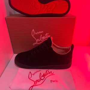 Svarta sneakers från Christian Louboutin - Snygga svarta sneakers från Christian Louboutin i mocka med klassisk röd sula och rund tå. Skorna har svarta snören och diskret branding på insidan. Perfekta för dig som vill ha en lyxig och stilren look med en ikonisk touch, och skrona är omvända alls helt ny med storlek 41 men passar också 42, jag fick vill sälja dem för att jag behöver inte ha dem.