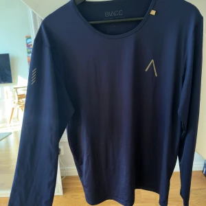 Mörkblå långärmad träningströja BLACC - Säljer en mörkblå långärmad träningströja från BLACC i storlek M. Tröjan är tillverkad i ett stretchigt material av polyester och elastan, har rund halsringning och diskreta tryckdetaljer på bröstet och ärmen. 