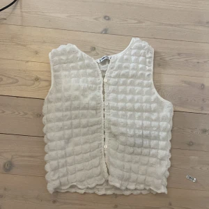 Vit quiltad väst från FB Sister - Superfin vit väst från FB Sister i quiltad stil med mjuk struktur och vågig kant. Västen har knäppning framtill med små knappar och är ärmlös. Perfekt att slänga över en långärmad tröja för en cool lager-på-lager-look.
