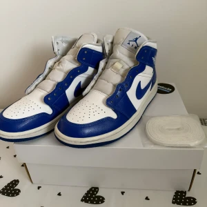 Nike Air Jordan 1 blå - Nike Air Jordan 1 sneakers i blått och vitt skinn med klassisk hög siluett. Snygg blå yttersula, perforerad tå och ikonisk swoosh på sidan. Kommer med extra vita skosnören. Perfekta för dig som vill sticka ut med en retro basketstil. Köpta från restocks. 