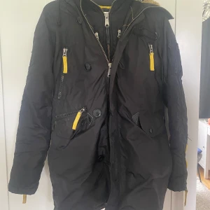 Svart/gul alpha industries jacka  - En svart alpha industries jacka med gula detaljer. Den har många dragkedjor och fickor, även fina detaljer. Luvan har päls inuti och en dragkedja. Jackan har små fläckar inuti, men det är inget som syns när man har på sig den 