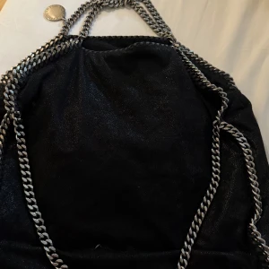 Svart axelväska Stella McCartney - Säljer en svart axelväska från Stella McCartney med kedjedetaljer i silver och glansig finish. Väskan har ett rosa innerfoder med logotypmönster och dragkedja på insidan. Perfekt för dig som gillar statement-accessoarer och snygga detaljer. Bra skick! Perfekt för skolan, då man får plats med dator+annat. Skriv för fler bilder💗nypris runt 10 000
