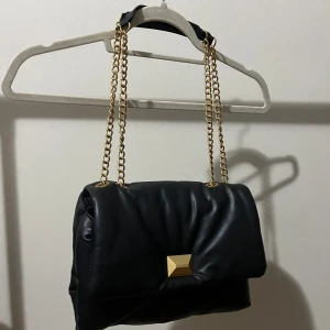 Svart axelväska med guldkedja från H&M - Snygg svart axelväska från H&M i mjukt syntetmaterial. Väskan har en guldfärgad kedja som axelrem och en dekorativ guldfärgad detalj på framsidan. Invändigt finns ett fack med dragkedja för småsaker. Perfekt för att lyfta din outfit.