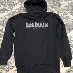 Svart hoodie från Balmain Paris  - Svart hoodie från Balmain Paris med stor logga framtill i vitt och rött. Klassisk känguruficka, ribbade muddar och huva. Mjuk insida och normal passform, perfekt för en stilren streetlook.