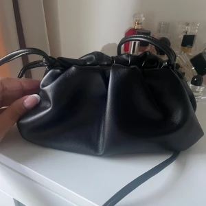 Svart dumpling väska i skinnimitation - Snygg svart axelväska ifrån Raffinato med trendig rynkad design och dragsko upptill. Väskan har ett mjukt, slätt material och smala axelband som knyts i sidorna. Perfekt storlek för det viktigaste och passar till många olika outfits.