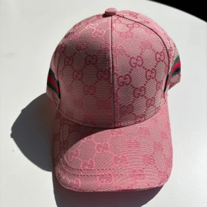 Rosa Gucci keps med GG-mönster - Snygg rosa keps från Gucci med klassiskt GG-monogram över hela kepsen. Har gröna och röda ränder på sidorna som ger extra lyxig känsla. Tillverkad i bomull och polyester, perfekt för dig som vill sticka ut med designerstil.