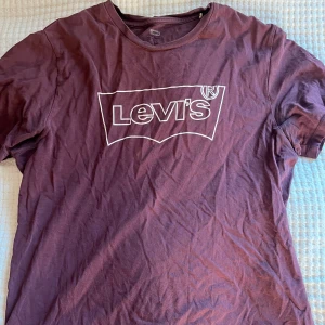 Vinröd Levi's t-shirt med logga - Snygg vinröd t-shirt från Levi's med klassisk vit logga tryckt på bröstet. Modellen är kortärmad och har rund hals. Materialet är mjuk bomull som känns skönt mot huden. Perfekt för dig som gillar streetwear och vill ha en enkel men ikonisk look.