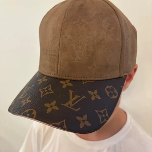 Keps i mocka och skinn - Snygg keps med brun mocka på ovansidan och klassisk monogrammönstrad skinnskärm i mörkbrunt och guld. Justerbar rem bak med samma monogramdetaljer. Perfekt för dig som gillar lyxiga accessoarer med statement.