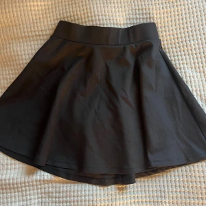 Svart kortkjol med utsvängd passform - Enkel och stilren svart kortkjol med bred midja och utsvängd modell. Kjolen är i ett mjukt och följsamt material som ger snyggt fall. Perfekt för dig som gillar en klassisk look med lite edge. Storlek 158/164 men passar xs-S