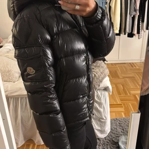 Svart Moncler Maya pufferjacka med huva - Säljer en svart pufferjacka från Moncler med glansig finish och huva. Jackan har dragkedja framtill, ficka med lock på ärmen och klassisk Moncler-logga. Perfekt för kalla dagar och riktigt snygg till streetwear-stilen. Pris kan diskuteras, köptes på Plick för 8k!💕