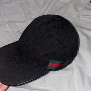 Svart keps från Gucci med logomönster - Säljer en svart keps från Gucci med klassiskt GG-logomönster över hela kepsen. Kepsen har justerbar rem baktill och en ikonisk grön och röd rand på sidan. Tillverkad i bomull och polyester, perfekt för dig som vill ha en stilren och lyxig accessoar. Pris kan diskuteras 