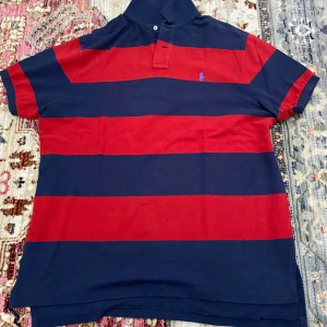 Randig röd och marinblå Polo t-shirt - Snygg randig t-shirt från Polo Ralph Lauren i rött och marinblått. Klassisk pikékrage med två knappar och broderad blå logga på bröstet. Tjocka horisontella ränder och korta ärmar.