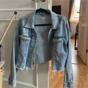 Croppad ljusblå jeansjacka - Säljer en croppad ljusblå jeansjacka med råa, fransiga kanter och klassisk krage. Jackan har två bröstfickor med lock och knappar samt långa ärmar. Perfekt för dig som vill ha en chill och trendig look.