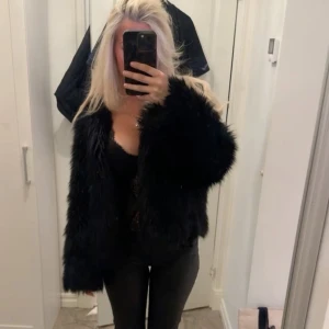 Svart pälsjacka med lång ärm - Säljer en svart pälsjacka med fluffig känsla och lång ärm. Jackan har en klassisk rak passform och är perfekt för dig som vill sticka ut med en trendig look. Ingen synlig dragkedja eller knappar framtill. Märke ej synligt.