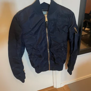 Mörkblå bomberjacka Alpha Industries - Snygg mörkblå bomberjacka från Alpha Industries med klassisk guldfärgad dragkedja och ärmficka med tygband. Jackan har ribbade muddar och är tillverkad i slitstark polyester. Perfekt för dig som vill ha en stilren och tidlös look.
