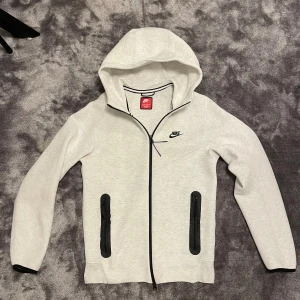 Nike tech vit - Nike hoodie i vitt med svart logga på bröstet. Tröjan har hel dragkedja, två svarta fickor med dragkedja och en stor huva. Perfekt för chill eller träning, riktigt clean och sportig look.
