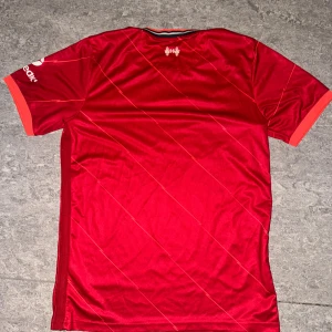 sushi -  KAN GÅ NER I SNABBT KÖP. Liverpool FCs officiella matchtröja i rött med tunna diagonala linjer, vita loggor och sponsortryck. Tillverkad i lätt och ventilerande polyester. Klassisk passform med korta ärmar och orange detaljer vid krage och ärmslut. Perfekt för dig som älskar fotboll och vill bära klubbens färger. 