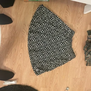 Svart blommig kortkjol med resår - Supersöt svart kortkjol med vita små blommor och bekväm resår i midjan. Kjolen har en lätt utställd form och är perfekt för dig som gillar en feminin och trendig stil. Passar perfekt till sneakers eller boots.