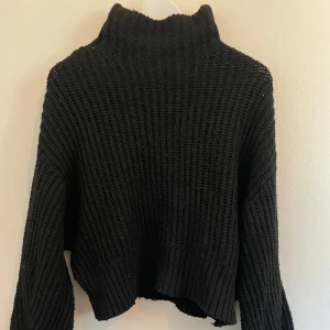 Svart stickad polotröja - Mysig svart stickad polotröja med grov ribbad struktur och hög krage. Tröjan har en något boxig och croppad passform med långa ärmar och breda muddar. Perfekt för kyliga dagar när du vill vara både varm och snygg.