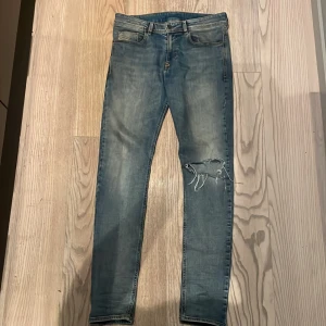 Blå jeans från Diesel - Snygga blå jeans från Diesel med slitningar och hål på ena benet för en cool look. Modellen är skinny fit med klassiska fem fickor och läderpatch bak i midjan. Perfekta för dig som gillar en trendig och avslappnad stil.