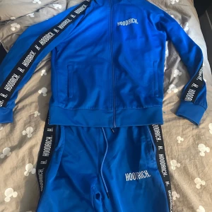 Blå hoodie från Hoodrich med dragkedja - Säljer en blå hoodie från Hoodrich med hel dragkedja och vita logotexter längs ärmarna. Tröjan har ribbade muddar, fickor på sidorna och svart detaljband med Hoodrich-tryck. Perfekt för en sportig och avslappnad stil.