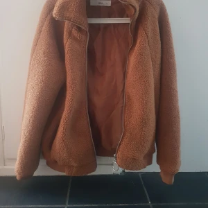 Brun teddyjacka från Ellos -  brun teddyjacka från Ellos med dragkedja och hög krage. Jackan har en oversized passform och är tillverkad i fluffigt teddy-material. 