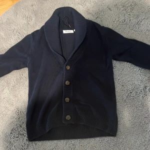 Mörkblå kofta från RYLS med knappar - Mysig mörkblå kofta  med bred sjalkrage och fyra stora knappar framtill. Tröjan är ribbstickad vid krage, knappslå och muddar. Perfekt för lager-på-lager och passar till många olika outfits. Parfeck nu till höst/vinter (kan diskutera pris)