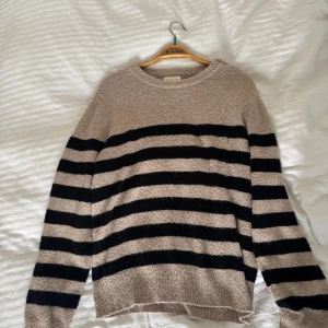 Randig stickad tröja med guldknappar - Beige och svart randig stickad tröja med snygga guldknappar på ena axeln. Tröjan har rund halsringning och långa ärmar. Perfekt för dig som gillar en chill men ändå stilren look.