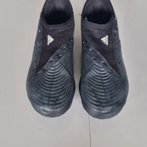 Svarta adidas Predator fotbollsskor storlek 34. - Säljer ett par svarta adidas Predator fotbollsskor med blå detaljer på dobbarna. Skorna har en strumpliknande ovandel i syntetmaterial och en mönstrad yta för extra grepp på bollen. Perfekta för dig som vill dominera på planen.
