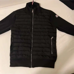 Svart Moncler cardigan herr - Svart pufferjacka från Moncler med quiltad front, höga krage och två dragkedjefickor. Jackan har Moncler-logga på ärmen och hel dragkedja framtill. Perfekt för dig som vill ha en stilren och sportig look med premiumkänsla.