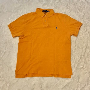 Persikofärgad pikétröja från Polo Ralph Lauren M - Persikofärgad pikétröja från Polo Ralph Lauren i bomull, custom slim fit. Klassisk krage med två knappar och blå broderad logga på bröstet. Perfekt för dig som gillar stilren och sportig vibe.