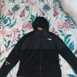 Svart vindjacka från The North Face - Säljer en svart vindjacka från The North Face med huva och hel dragkedja. Jackan har en stilren design med logga på bröstet och är tillverkad i lätt polyester. Perfekt för dig som gillar sportig och clean stil.