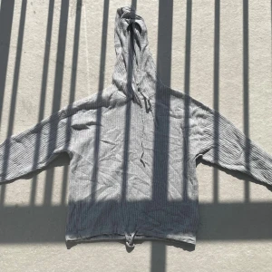 Grå ribbad hoodie med snörning - Grå ribbad hoodie med huva och snörning både i halsen och nedtill. Tröjan har långa ärmar och är tillverkad i ett mjukt, stickat material som känns skönt mot huden. Perfekt för dig som gillar en avslappnad och stilren look.