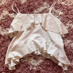 Vit volangtop från Nelly XS - Supersöt vit linnetopp från Nelly i storlek XS. Toppen har tunna axelband och är dekorerad med volanger både upptill och nedtill. Materialet är lätt strukturerat och ger en snygg textur. Perfekt för varma dagar när du vill ha en luftig och trendig look.