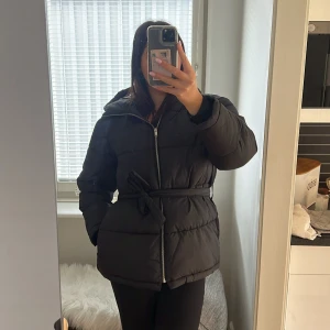 Svart pufferjacka från NLY Trend - Svart pufferjacka från NLY Trend med luva, dragkedja och bälte i midjan. Och är tillverkad i syntetmaterial. Perfekt för kalla dagar och har en snygg, lite oversized passform. Jackan är endast använd 1-2 gånger.