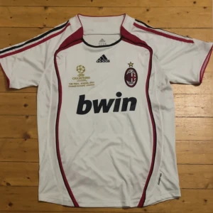 AC Milan Kaka Champions League 2007 borta tröja  - Säljer en vit AC Milan fotbollströja från Adidas med röda och svarta detaljer, Champions League-finaltryck och Kaka 22 på ryggen. Tröjan har korta ärmar, klubbmärke på bröstet och Bwin som sponsor. Materialet är lätt polyester, perfekt för match eller träning.