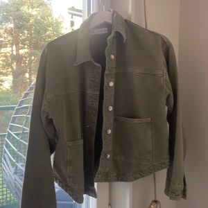 Olivgrön jeansjacka från Zara - Säljer en olivgrön jeansjacka från Zara i storlek S. Jackan har klassisk krage, silverfärgade knappar framtill och två stora fickor. Perfekt att slänga på sig över en hoodie eller t-shirt. Enkel och stilren design som funkar till det mesta.