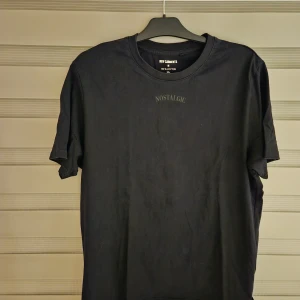 Svart t-shirt från New Garments - Svart t-shirt från New Garments i 100% bomull med trycket 'NOSTALGIC' på bröstet. Klassisk rund halsringning och korta ärmar. Enkel och stilren design som passar till allt.